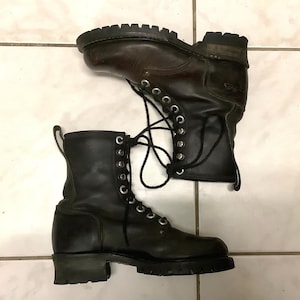 May include: Two black leather combat boots with dark brown detailing. The boots feature a lace-up design with silver eyelets and chunky, lug soles. The boots are positioned on a tiled surface.