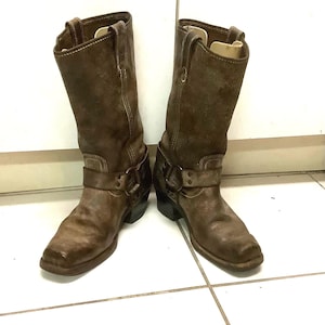 Puede incluir: Un par de botas de vaquero de cuero marrón con un aspecto desgastado. Las botas tienen punta cuadrada, caña media y un detalle de hebilla alrededor del tobillo. Las botas están sobre un fondo blanco.