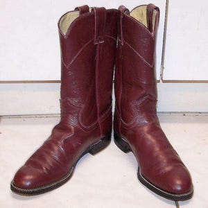 Könnte beinhalten: Ein Paar dunkelburgunderfarbene Leder-Cowboystiefel mit abgerundeter Zehenpartie, einem wadenhohen Schaft mit seitlichen Anziehschlaufen und einem Blockabsatz. Die Stiefel haben sichtbare Ziernähte am Vorderblatt und Schaft sowie eine braune Sohle.