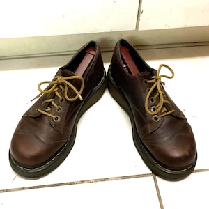 Può includere: Un paio di scarpe Oxford in pelle marrone con lacci gialli. Le scarpe hanno una punta arrotondata e una suola spessa nera. Le scarpe sono esposte su una superficie piastrellata bianca.