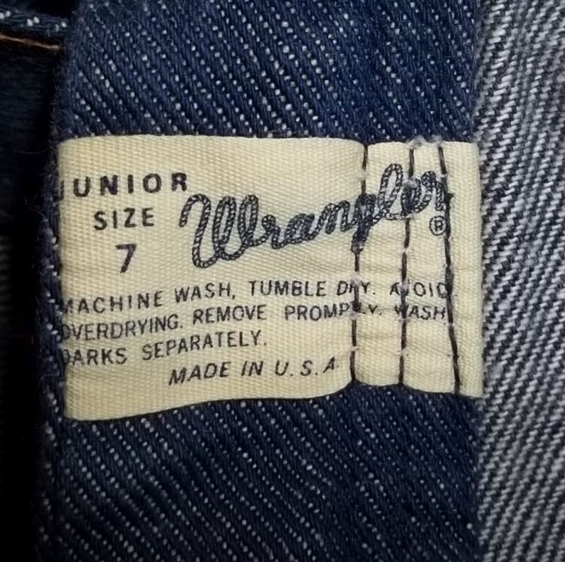 Peut inclure: Gros plan sur un tissu denim avec une &eacute;tiquette rectangulaire. L'&eacute;tiquette indique "JUNIOR SIZE 7 Wrangler" et comprend des instructions de lavage. L'&eacute;tiquette est de couleur cr&egrave;me avec du texte noir. "MADE IN U.S.A." est imprim&eacute; en bas.