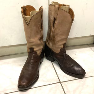 May include: A pair of two-toned cowboy boots featuring dark brown textured leather vamps and shafts with lighter tan leather uppers. The boots have a pointed toe, a stacked heel, and pull tabs at the top of the shafts, with internal inserts visible.