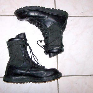 Puede incluir: Dos botas de combate de cuero negro con diseño de cordones. Las botas tienen un estilo de caña alta con un panel de tela texturizada en los laterales y una suela gruesa y resistente. Las botas están diseñadas para la durabilidad y la protección.