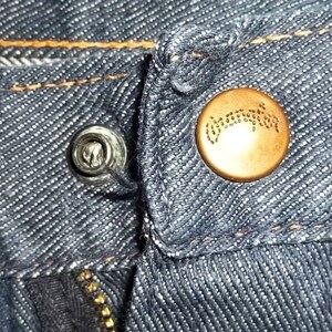 Peut inclure: Gros plan sur un jean en denim montrant un rivet argent&eacute; et un bouton de couleur cuivre. Le bouton a un motif en relief. Le jean a des coutures visibles et une fermeture &eacute;clair. Le tissu est de couleur bleu fonc&eacute;.