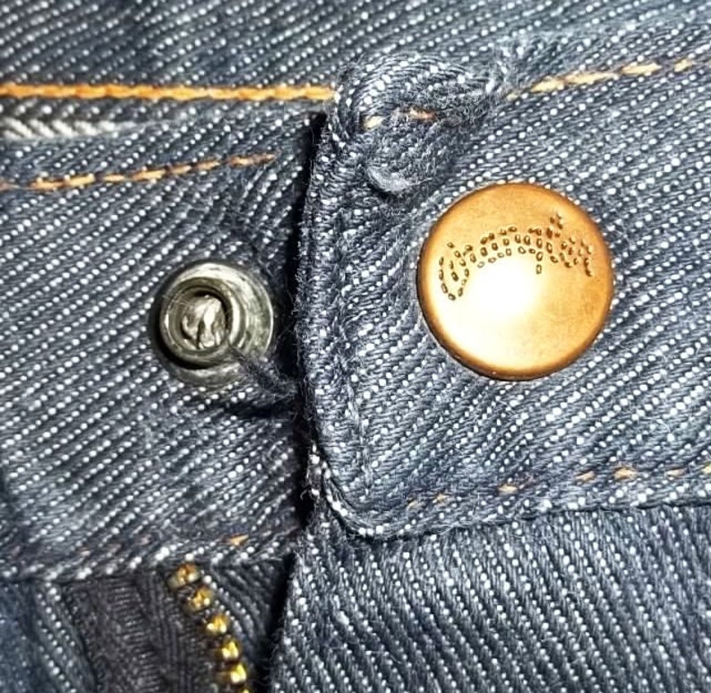 Peut inclure: Gros plan sur un jean en denim montrant un rivet argent&eacute; et un bouton de couleur cuivre. Le bouton a un motif en relief. Le jean a des coutures visibles et une fermeture &eacute;clair. Le tissu est de couleur bleu fonc&eacute;.