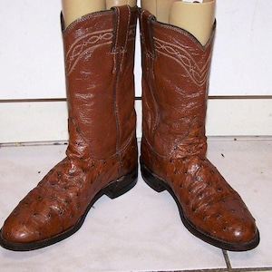 Op de afbeelding: Een paar bruine leren cowboylaarzen met een textuurpatroon op de voet en een gladde leren schacht met decoratieve witte stiksels langs de bovenrand. De laarzen hebben een ronde neus, een lage hak en een donkerbruine zool.