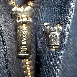 Peut inclure: Gros plan sur une fermeture &eacute;clair en laiton sur un tissu denim bleu fonc&eacute;. Le curseur de la fermeture &eacute;clair porte la marque "SCOVILL". La fermeture &eacute;clair est partiellement ferm&eacute;e, r&eacute;v&eacute;lant les dents et la texture du tissu.