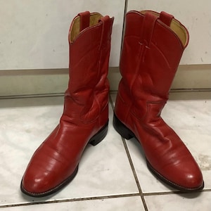 Op de afbeelding: Een paar felrode cowboylaarzen. De laarzen hebben een klassiek western design met een puntige neus, een middelhoge schacht en een gestapelde hak. Ze zijn gemaakt van een glad, glanzend materiaal, waarschijnlijk leer.