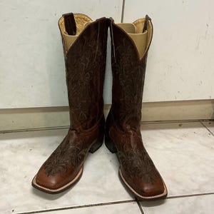 May include: A pair of dark brown leather cowboy boots featuring an intricate embossed floral pattern across the shaft and foot. These boots have a square toe design, a stacked heel, and light-colored interior lining with visible boot inserts.