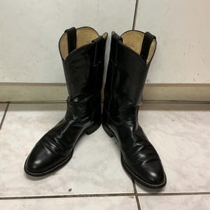 Könnte beinhalten: Ein Paar schwarze Leder-Cowboystiefel mit poliertem Finish, spitzer Zehenpartie, wadenhohem Schaft und Zuglaschen am oberen Rand. Das Innenfutter ist hellbeige, und die Stiefel haben einen niedrigen, geschichteten Absatz sowie sichtbare Nähte.