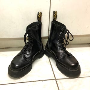 Könnte beinhalten: Ein Paar schwarze Lederstiefeletten mit dicken Plateausohlen, ausgestattet mit schwarzen Schnürsenkeln, silberfarbenen Ösen und einem seitlichen Reißverschluss. Jeder Stiefel hat eine gelb-schwarze AirWair-Zugschlaufe und sichtbare gelbe Nähte.