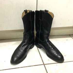 Könnte beinhalten: Ein Paar schwarze Leder-Cowboystiefel. Die Stiefel haben ein klassisches Design mit spitzer Zehenpartie und einem mittelhohen Schaft. Das Leder scheint glatt und gut gepflegt zu sein, mit subtilen Knitterfalten. Die Stiefel stehen vor einem neutralen Hintergrund.