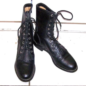 Könnte beinhalten: Ein Paar schwarze Schnürstiefel aus Leder mit abgerundeter Zehenpartie, silberfarbenen Metallösen und einem dekorativen Fransen-Kiltie-Einsatz über dem Vorderfuß. Die Stiefel haben schwarze Schnürsenkel, eine glatte Oberfläche und eine mittlere Schafthöhe.