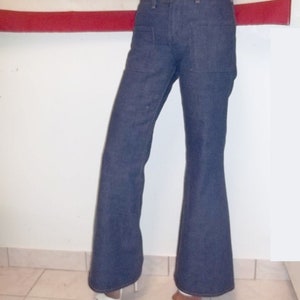 Peut inclure: Pantalon &eacute;vas&eacute; en denim bleu fonc&eacute;. Le pantalon a une taille haute et une jambe &eacute;vas&eacute;e. Le mod&egrave;le porte des sandales &agrave; talons hauts blanches. L'arri&egrave;re-plan comprend un drapeau rouge et blanc.