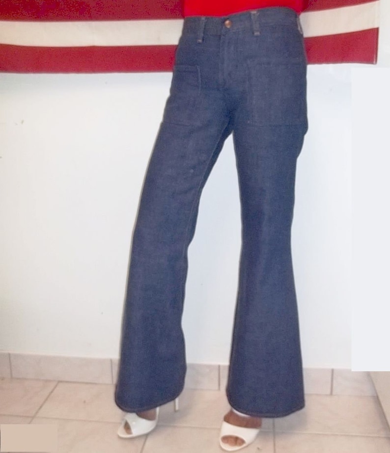 Peut inclure: Pantalon &eacute;vas&eacute; en denim bleu fonc&eacute;. Le pantalon a une taille haute et une jambe &eacute;vas&eacute;e. Le mod&egrave;le porte des sandales &agrave; talons hauts blanches. L'arri&egrave;re-plan comprend un drapeau rouge et blanc.