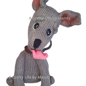 Puede incluir: Un perro tejido a mano de color gris con grandes ojos negros de botón, un collar marrón y una lengua rosa. El perro tiene orejas grandes y caídas con el interior rosa. El texto "A Crafty Life by Mandy" es visible.