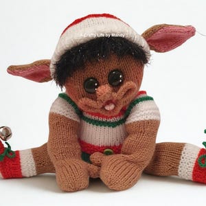 May include: Hand-knitted elf doll with a red and white striped hat, brown skin, and large black eyes. The elf wears a striped shirt with green and red accents. The doll's legs are outstretched, and the feet are adorned with bells.