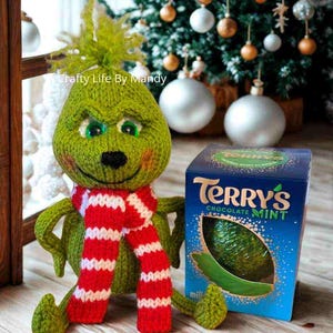 Puede incluir: Una muñeca Grinch verde hecha a mano con una bufanda a rayas rojas y blancas, junto a una caja de Terry's Chocolate Mint. El Grinch tiene una cara sonriente y está frente a un árbol de Navidad.