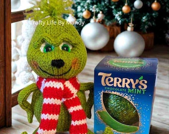 Może przedstawiać: Ręcznie robiona zielona lalka Grincha z czerwono-białym szalikiem, obok pudełka Terry's Chocolate Mint. Grinch ma uśmiechniętą twarz i siedzi przed udekorowaną choinką.