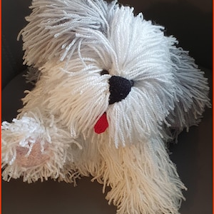May include: A handmade yarn dog plushie with white and gray fur. The dog has a black nose, a red tongue, and pink paws. The plushie is made of yarn and is designed to look like an Old English Sheepdog.