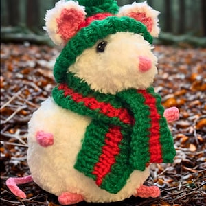 May include: A soft white mouse toy, adorned with a green and red striped scarf, a green hat with a bobble, and pink ears, nose, feet, and tail. The toy is set against a backdrop of autumn leaves and a blurred forest.