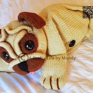 May include: A knitted plush toy pug dog in beige and brown with black eyes. The toy is lying on a white surface. "Crafty Life by Mandy" is written on the toy.