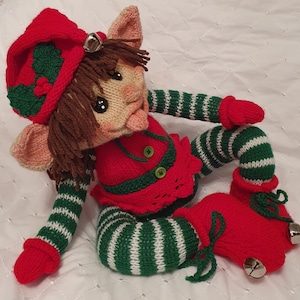 May include: A knitted Christmas elf doll. It has a red hat with green holly, a red outfit with green trim, and green and white striped legs and arms. The doll has a smiling face, brown hair, and bells on its shoes.