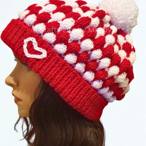 May include: A red and white crocheted beanie featuring a checkered puff stitch pattern, a solid red ribbed brim, a white embroidered heart outline on the side, and a large white yarn pom-pom on top. The text A Crafty Life by Mandy is visible.
