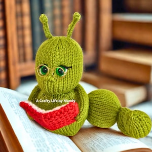 以下が含まれることがあります： 金縁のメガネをかけ、赤い本を読んでいる緑色の編み物で作られたイモムシの置物。 置物には大きな緑色の目と触角があります。「A Crafty Life by Mandy」の文字が見えます。