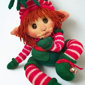 Op de afbeelding: Een gebreide elfenpop met rood haar, met een rood-wit gestreepte muts met een maretak en een belletje. De pop heeft een groen vestje, rood-wit gestreepte mouwen en leggings, en groene schoenen met rode veters en kleine belletjes.