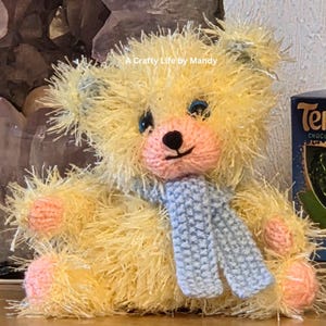 May include: A fluffy, handmade yellow teddy bear wearing a light blue scarf. The bear has a pink muzzle, paws, and ears, and blue eyes. The text "A Crafty Life by Mandy" is visible. A box of Terry's Chocolate is in the background.