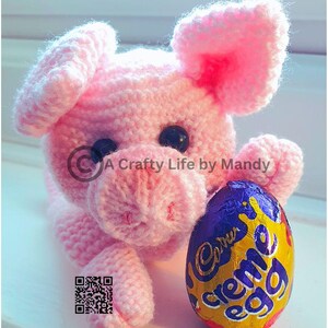 May include: A pink crocheted pig holding a Cadbury Creme Egg chocolate. The pig has a pink nose and black eyes. The egg is wrapped in gold foil with the Cadbury Creme Egg logo.