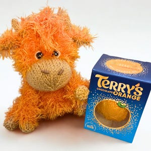 May include: An orange, shaggy, knitted Highland cow plush toy next to a blue box of Terry's Chocolate Orange. The box has gold text and a picture of an orange chocolate ball. The plush toy has a tan face and small, dark eyes.
