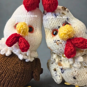 May include: Two handmade knitted chicken dolls. One chicken is white with a brown body, red comb and wattle, yellow beak and feet. The other chicken is off-white with a speckled pattern, red comb and wattle, yellow beak and feet. Text: A Crafty Life by Mandy.