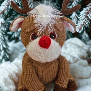 May include: A knitted reindeer toy in shades of brown and white, with a red nose and brown antlers. The text "A Crafty Life by Mandy" is visible. The reindeer is set against a snowy backdrop, evoking a festive atmosphere.