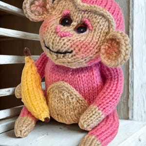 Könnte beinhalten: Eine handgestrickte Affenpuppe in Rosa und Beige, die eine gelbe Banane hält. Der Affe hat ein lächelndes Gesicht mit schwarzen Augen und beige Details an Gesicht, Ohren, Händen und Füßen. Die Puppe sitzt auf einer weißen Oberfläche.