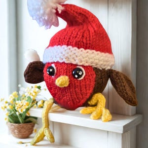 Könnte beinhalten: Handgestricktes Weihnachtsrotkehlchen mit roter Weihnachtsmütze und weißem Bommel. Der Vogel hat eine rote Brust, braune Flügel und gelbe Füße. Es sitzt auf einem weißen Regal, im Hintergrund eine kleine Topfpflanze.
