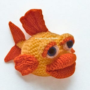 May include: A handmade crochet fish in shades of yellow and orange. The fish has large blue eyes, orange lips, and fins. The text "A Crafty Life by Mandy" is visible.