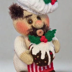 Op de afbeelding: Een gebreide pop in de vorm van een kok met een witte muts versierd met groene hulst en rode bessen, een witte jas en een rood schort. De pop heeft een bruine snor, roze wangen en houdt een donkerbruine kerstpudding met witte glazuur vast.