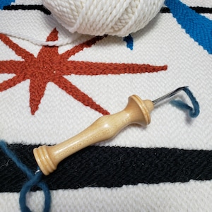 May include: A wooden tapestry needle with a dark blue yarn attached to the hook. The needle is lying on a white and black striped surface with a red star design.