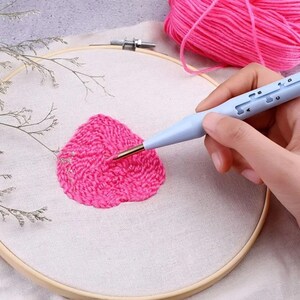 Puede incluir: Primer plano de una mano que utiliza una herramienta de punzón de aguja azul y dorada para crear un diseño de flor rosa en una tela blanca estirada en un aro de bordado de madera.