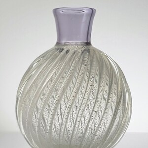 Pu&ograve; includere: Un vaso di vetro trasparente con un design a spirale e texture. Il vaso ha un bordo viola chiaro e un corpo rotondo e bulboso.
