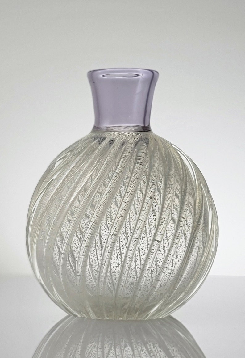 Pu&ograve; includere: Un vaso di vetro trasparente con un design a spirale e texture. Il vaso ha un bordo viola chiaro e un corpo rotondo e bulboso.
