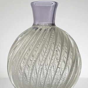 Pu&ograve; includere: Un vaso di vetro trasparente con un design a spirale e texture. Il vaso ha un corpo rotondo e un collo corto e largo. La parte superiore del collo &egrave; di colore lilla chiaro.