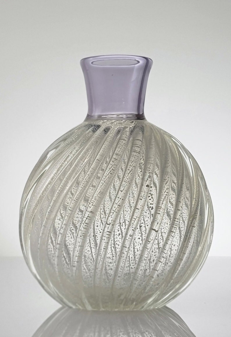 Pu&ograve; includere: Un vaso di vetro trasparente con un design a spirale e texture. Il vaso ha un corpo rotondo e un collo corto e largo. La parte superiore del collo &egrave; di colore lilla chiaro.