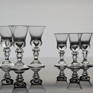 May include: Set of six clear glass shot glasses with a unique design. The glasses have a round base and a stem with a bulbous design. The glasses are arranged in two rows of three.