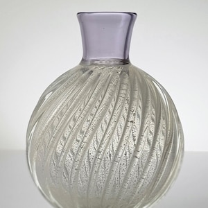Pu&ograve; includere: Un vaso di vetro trasparente con un design a spirale e texture. Il vaso ha un bordo di colore lilla chiaro e un corpo rotondo e bulboso.