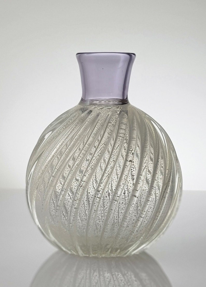Pu&ograve; includere: Un vaso di vetro trasparente con un design a spirale e texture. Il vaso ha un bordo di colore lilla chiaro e un corpo rotondo e bulboso.