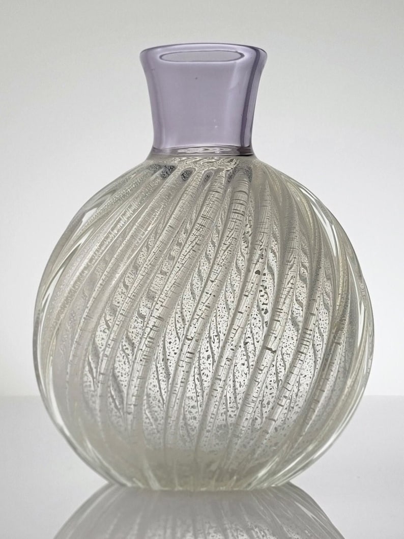Pu&ograve; includere: Un vaso di vetro trasparente con un design a spirale e texture e un bordo di colore lilla chiaro. Il vaso ha un corpo rotondo e un collo stretto.