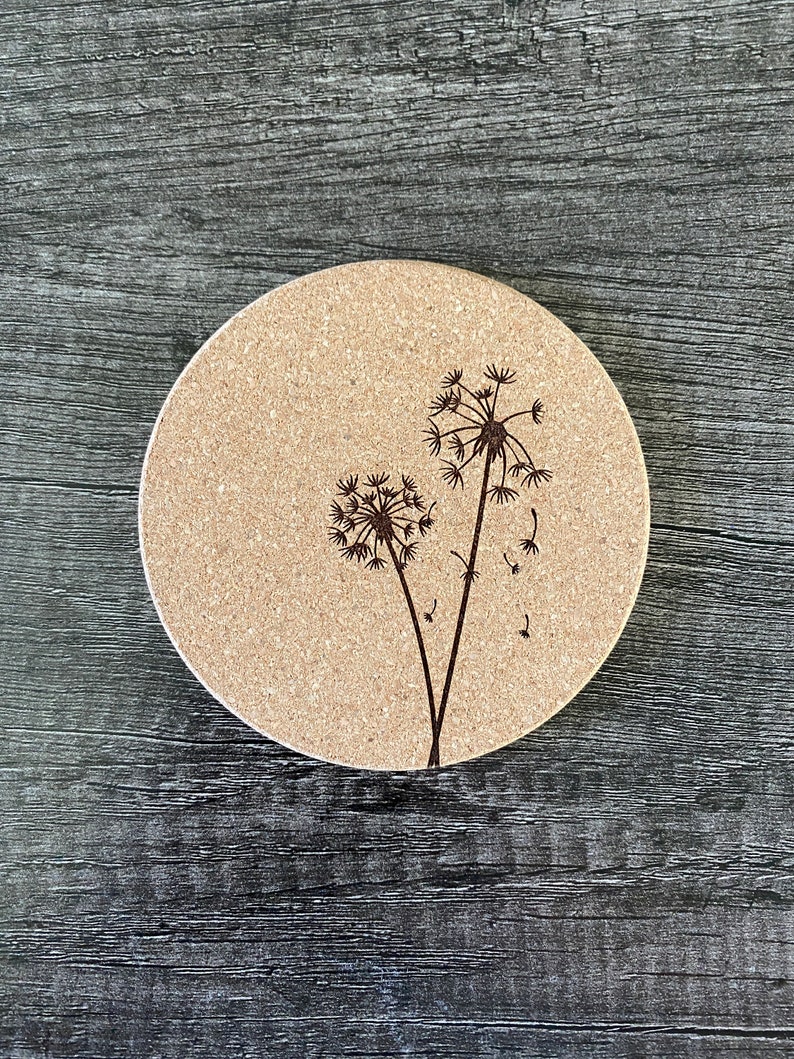 May include: A round cork coaster with a laser-etched design of two dandelions. The dandelions are black and the background is a light brown.